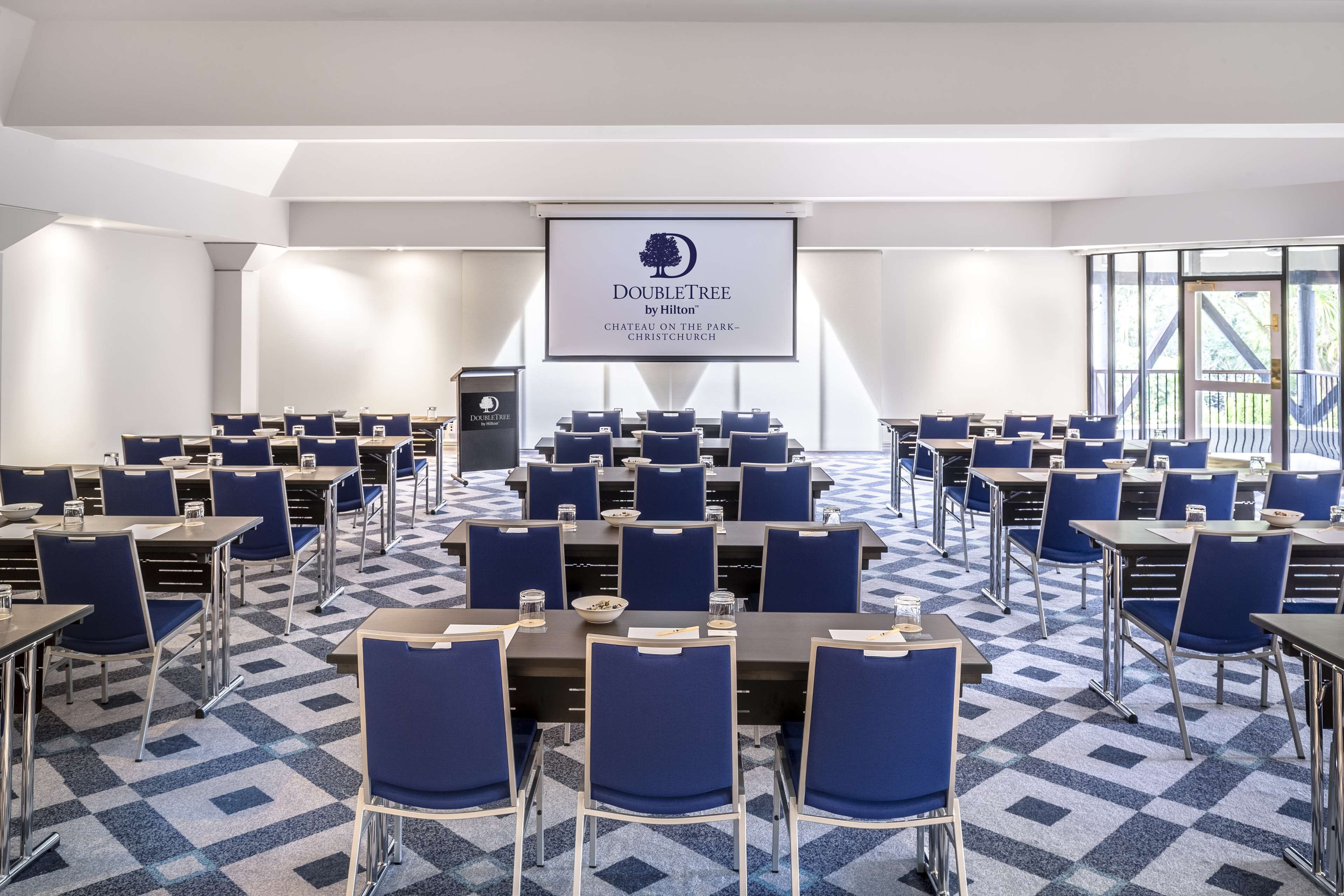 Sala de conferências com mesas e cadeiras dispostas para uma reunião. Uma tela exibe o logotipo e a localização DoubleTree.