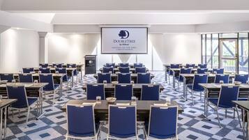 Sala de conferências com mesas e cadeiras dispostas para uma reunião. Uma tela exibe o logotipo e a localização DoubleTree.