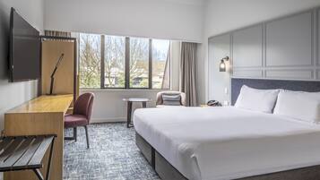 A hotel room with a large bed, a desk, a chair, a small table, a TV, and a window with a view of trees and buildings.