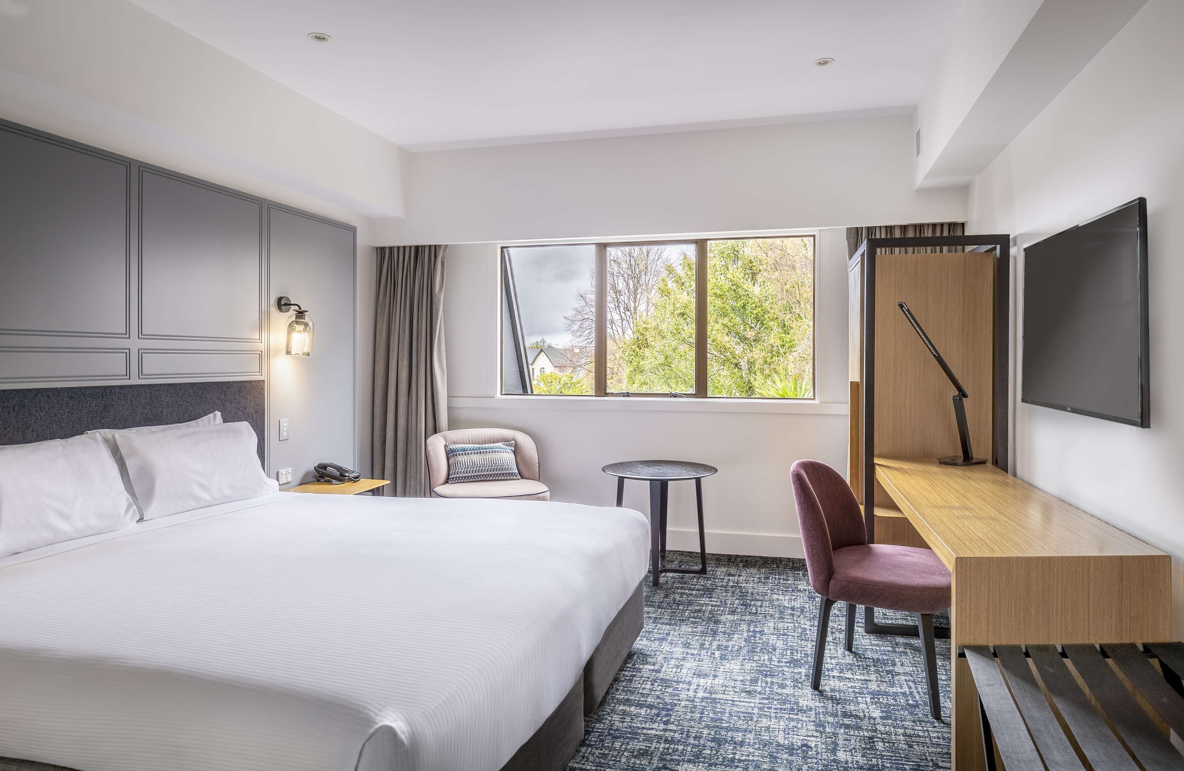A hotel room with a large bed, a desk, a chair, a small table, a TV, and a window with a view of trees.