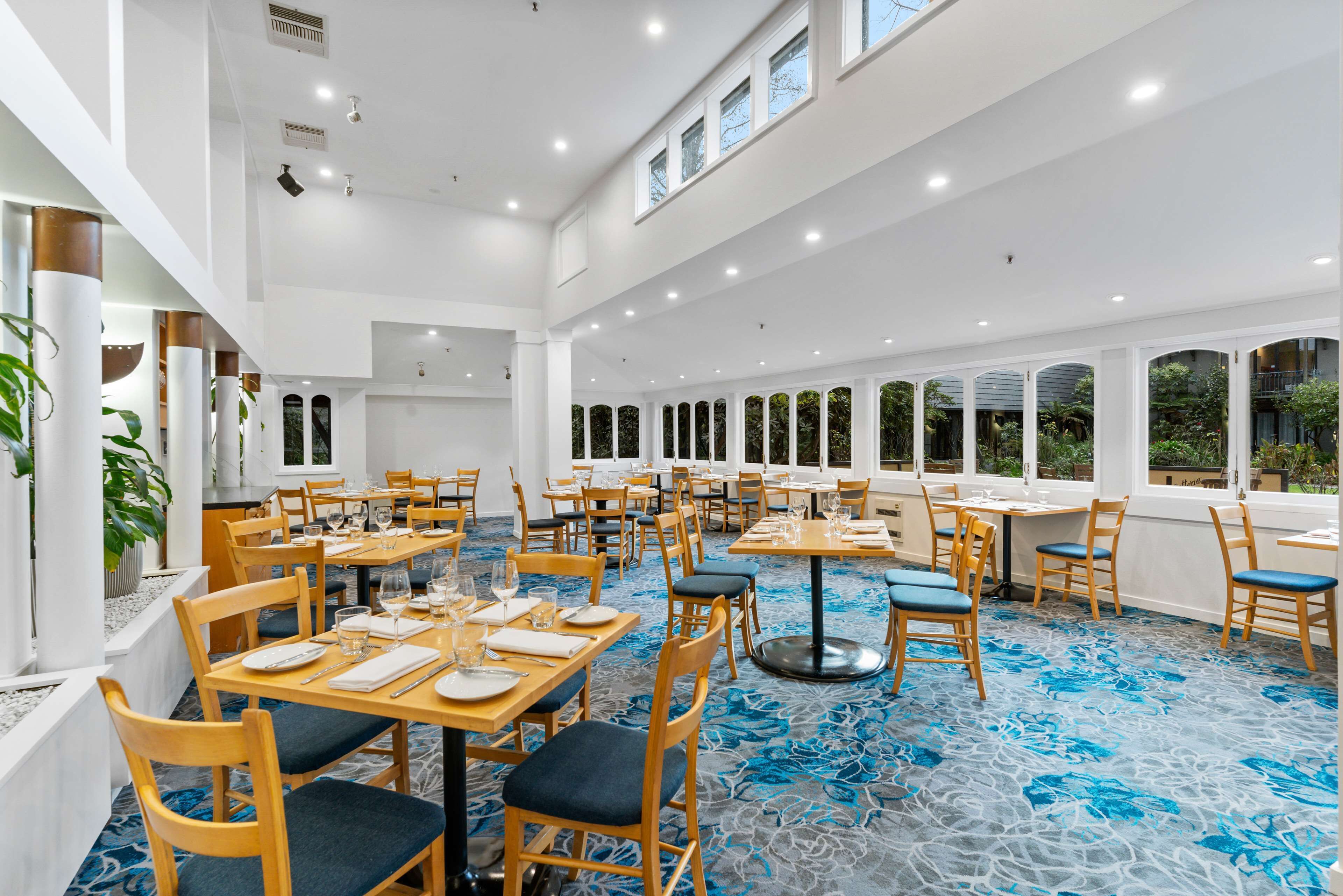 A spacious dining area with multiple tables and chairs, large windows, and a blue patterned carpet.