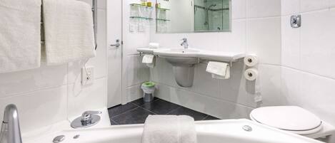 Un baño moderno con bañera, lavabo e inodoro. Toallas cuelgan de un perchero, recinto de ducha con mampara de vidrio y un espejo sobre el lavabo.