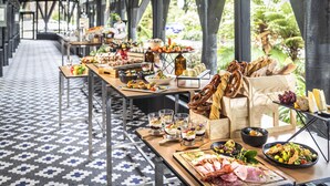 Una larga mesa con diversos alimentos y bebidas, ubicada en un espacio cubierto con piso estampado y grandes ventanales.