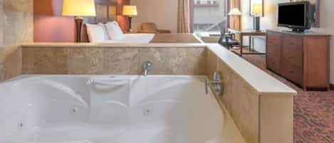 A hotel room with a large bathtub, a bed, a desk with a television, and a mirror.