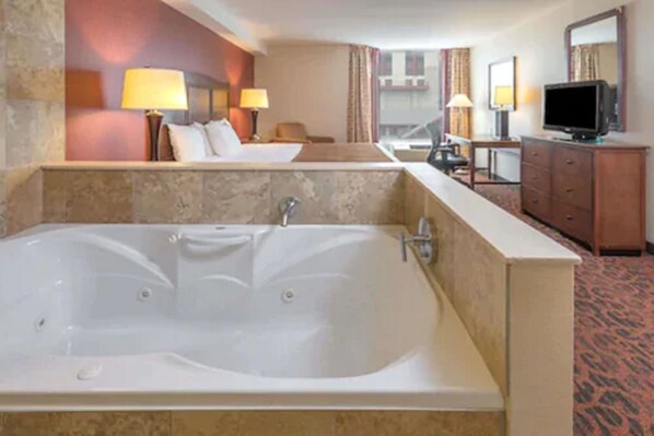 A hotel room with a large bathtub, a bed, a desk with a television, and a mirror.