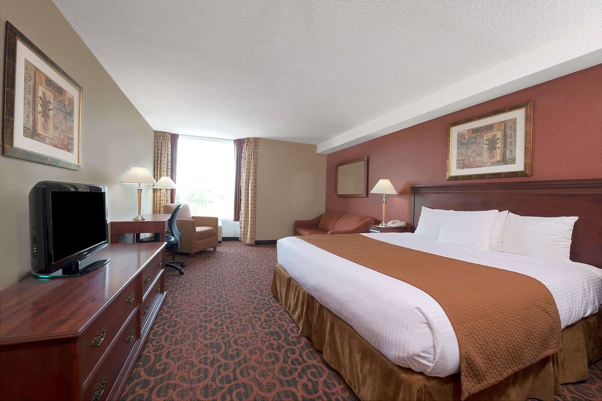 Habitación de hotel con cama, escritorio con televisor, silla, lámpara y ventana con cortinas.