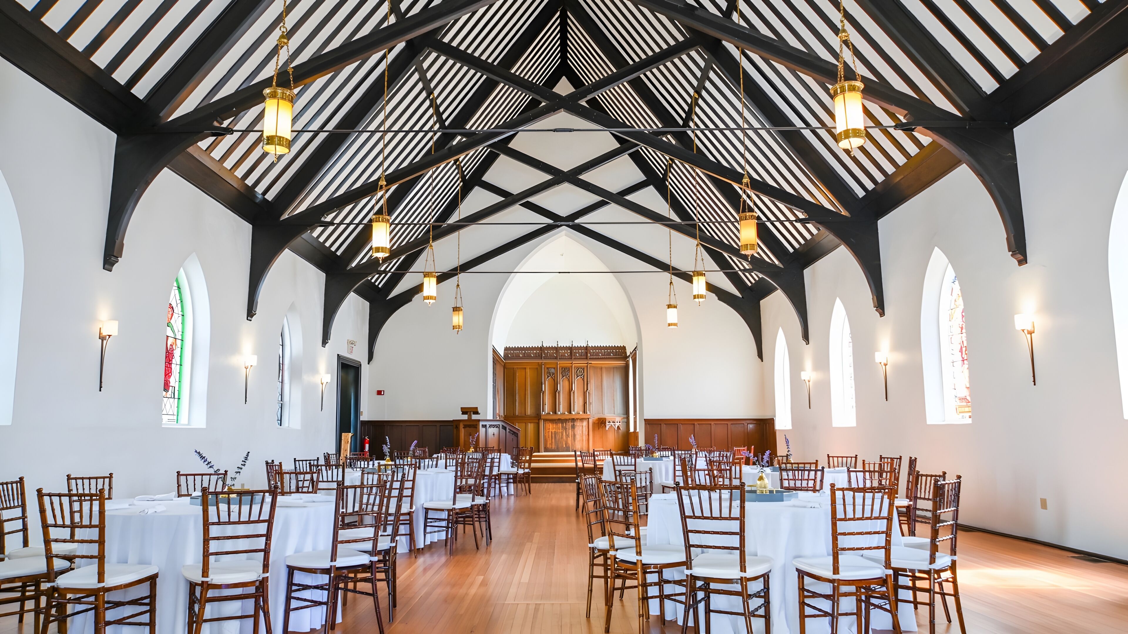 Un vaste hall doté de hauts plafonds, de poutres en bois et de nombreux lustres. Des tables sont disposées pour accueillir les invités, et de grandes fenêtres ornées de vitraux illuminent l’espace.