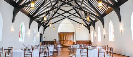 A spacious hall with high ceilings, wooden beams, and multiple chandeliers. Tables are arranged for seating, and there are large windows with stained glass.