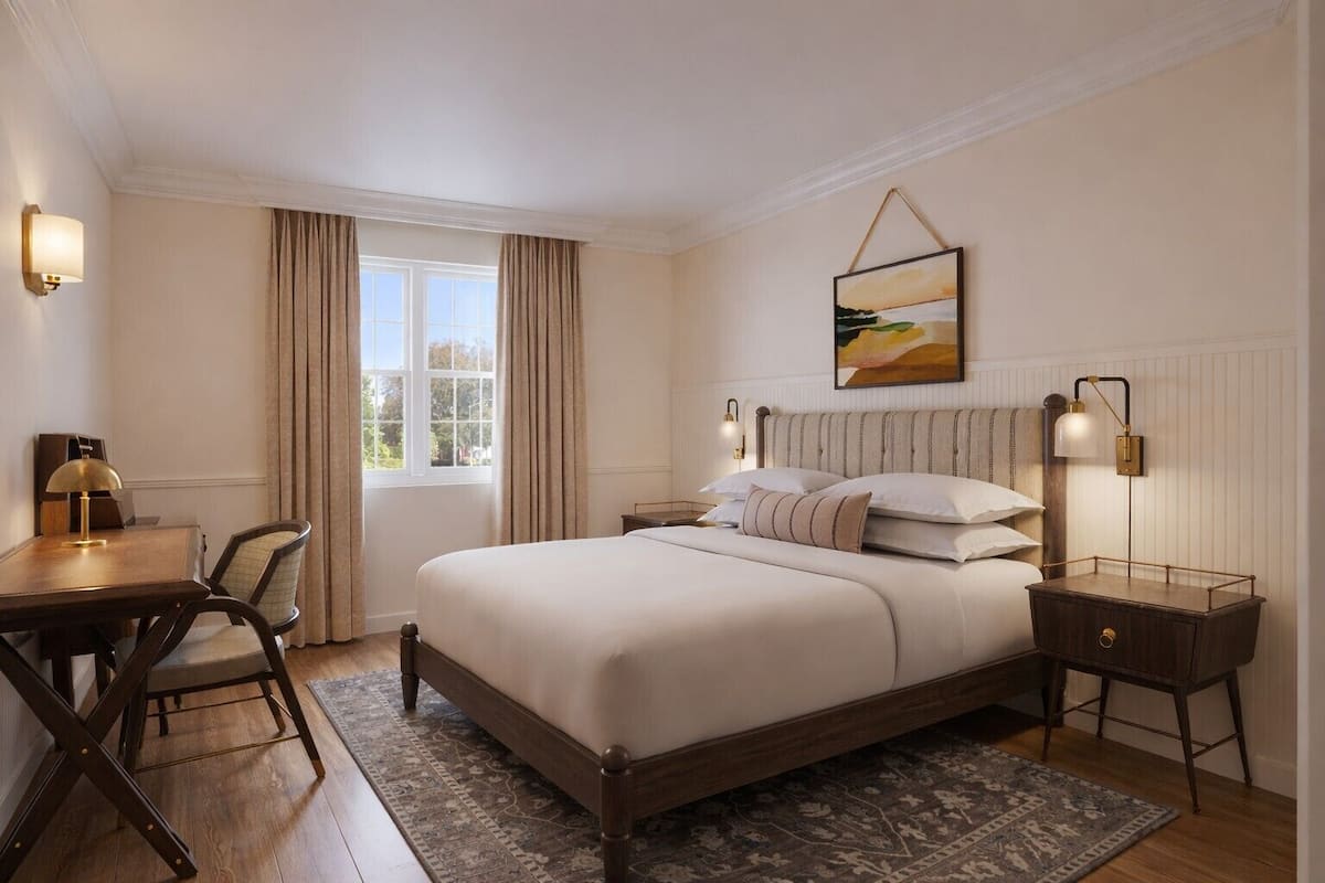 A hotel room with a bed, bedside tables, a desk, a chair, a window with curtains, and a painting on the wall.
