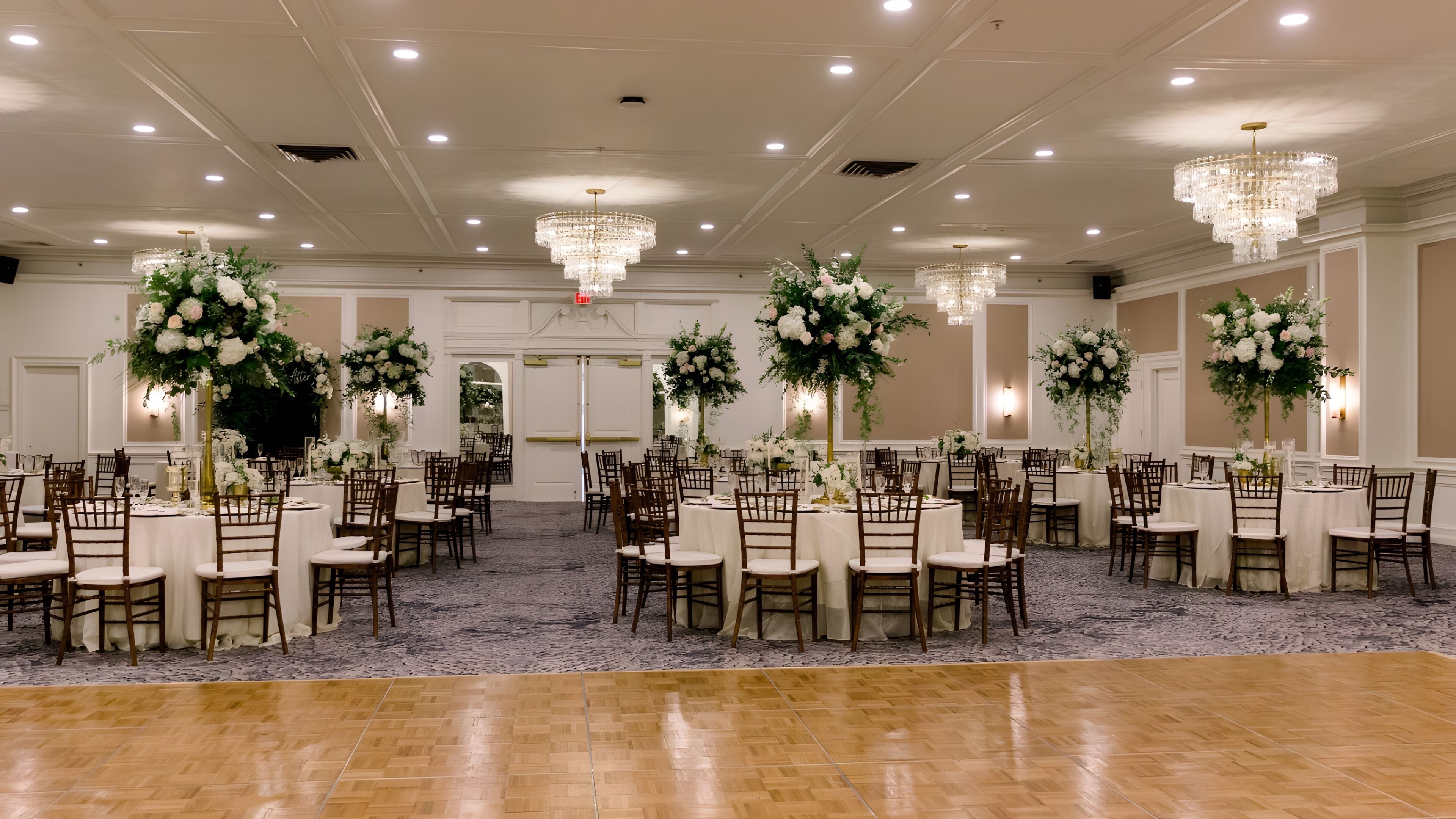 A banquet hall with round tables, wooden chairs, and floral centerpieces.