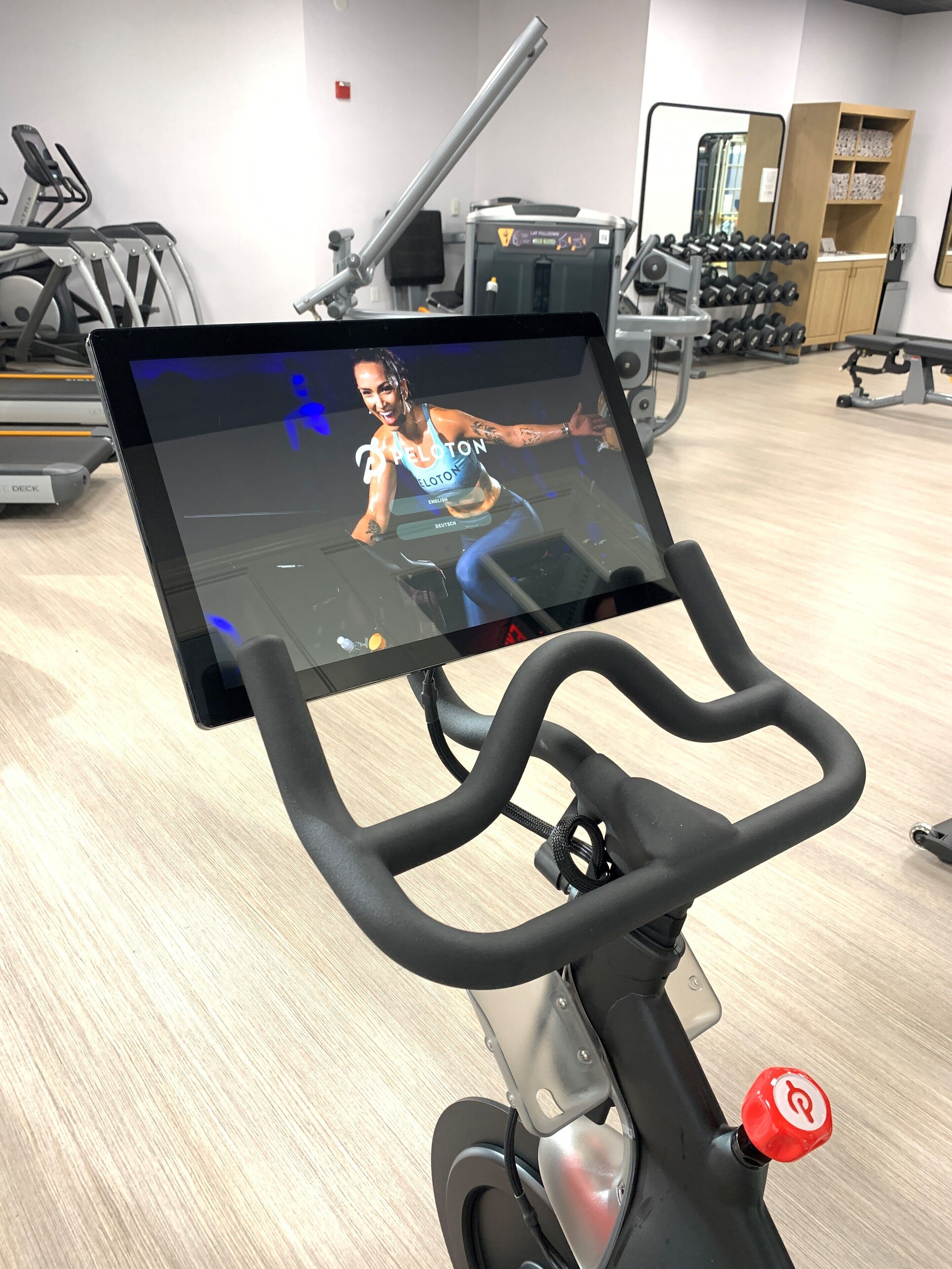 Una bicicleta estática con una pantalla digital mostrando un video de entrenamiento, rodeada de equipo de gimnasio.