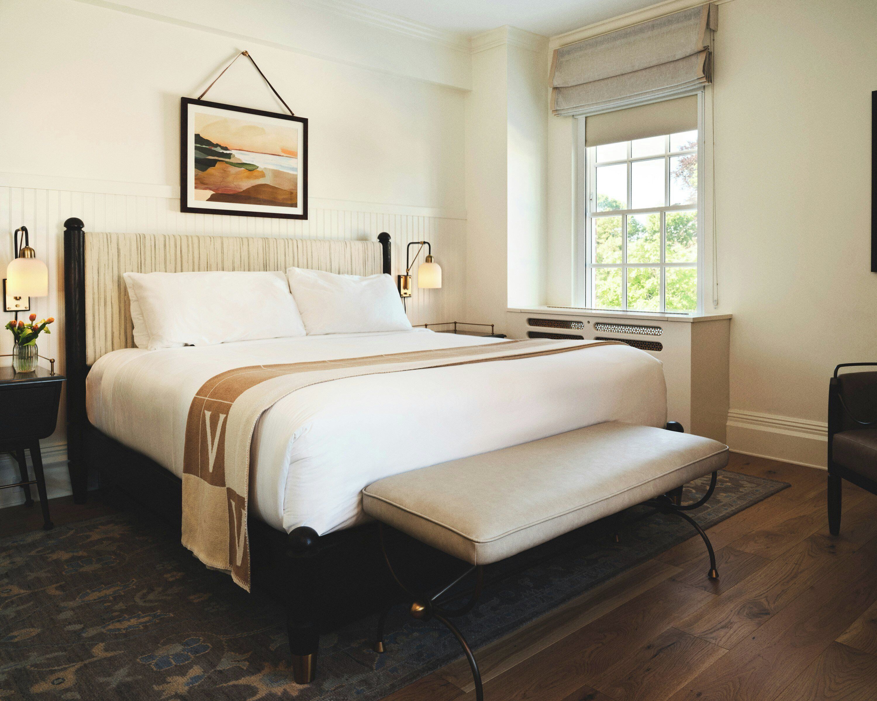A large bed with a bench, a bedside table with a lamp, a framed picture on the wall, and a window with a view of greenery.