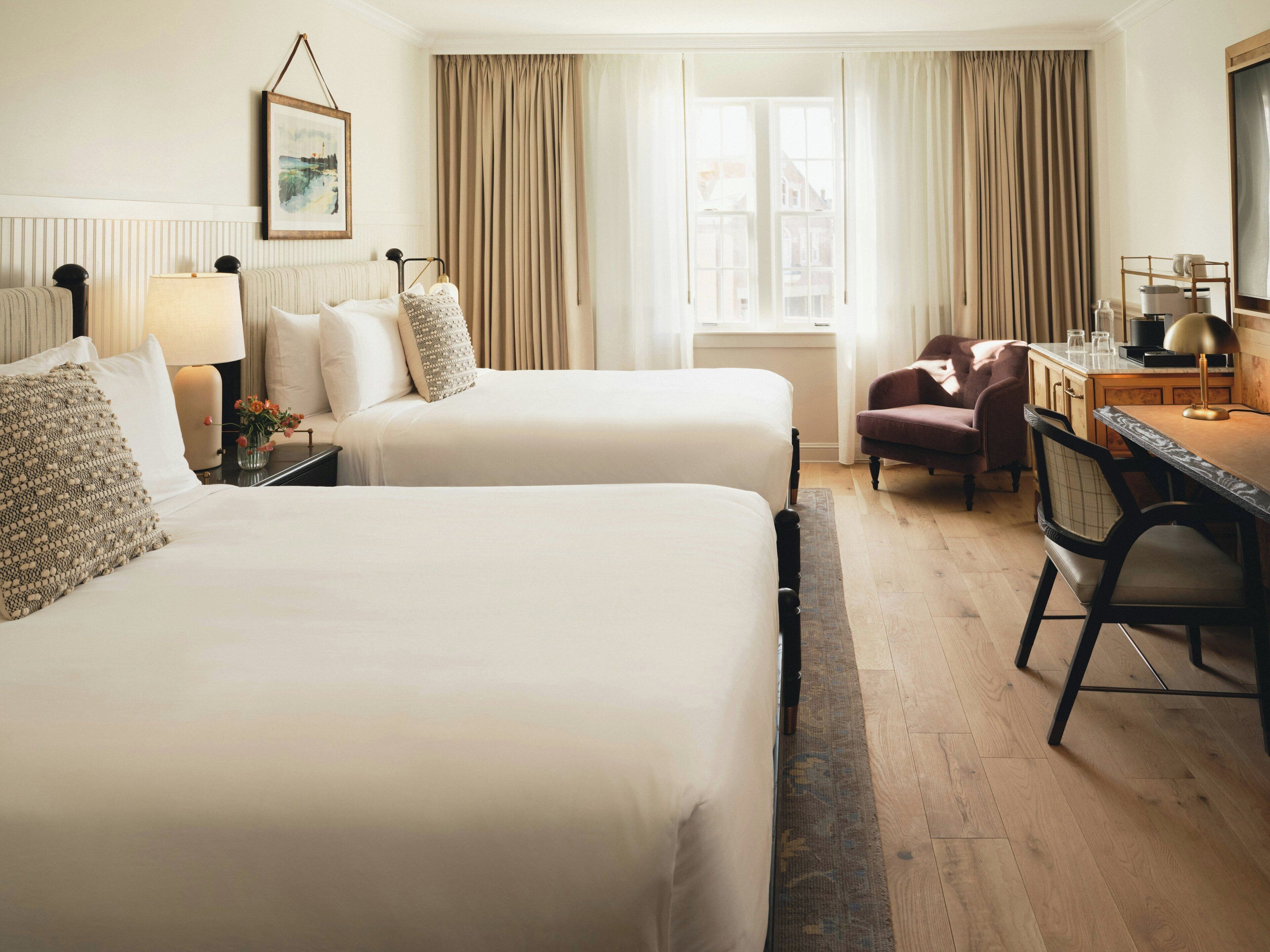 A hotel room with two beds, a desk, a chair, a lamp, and a window with curtains.