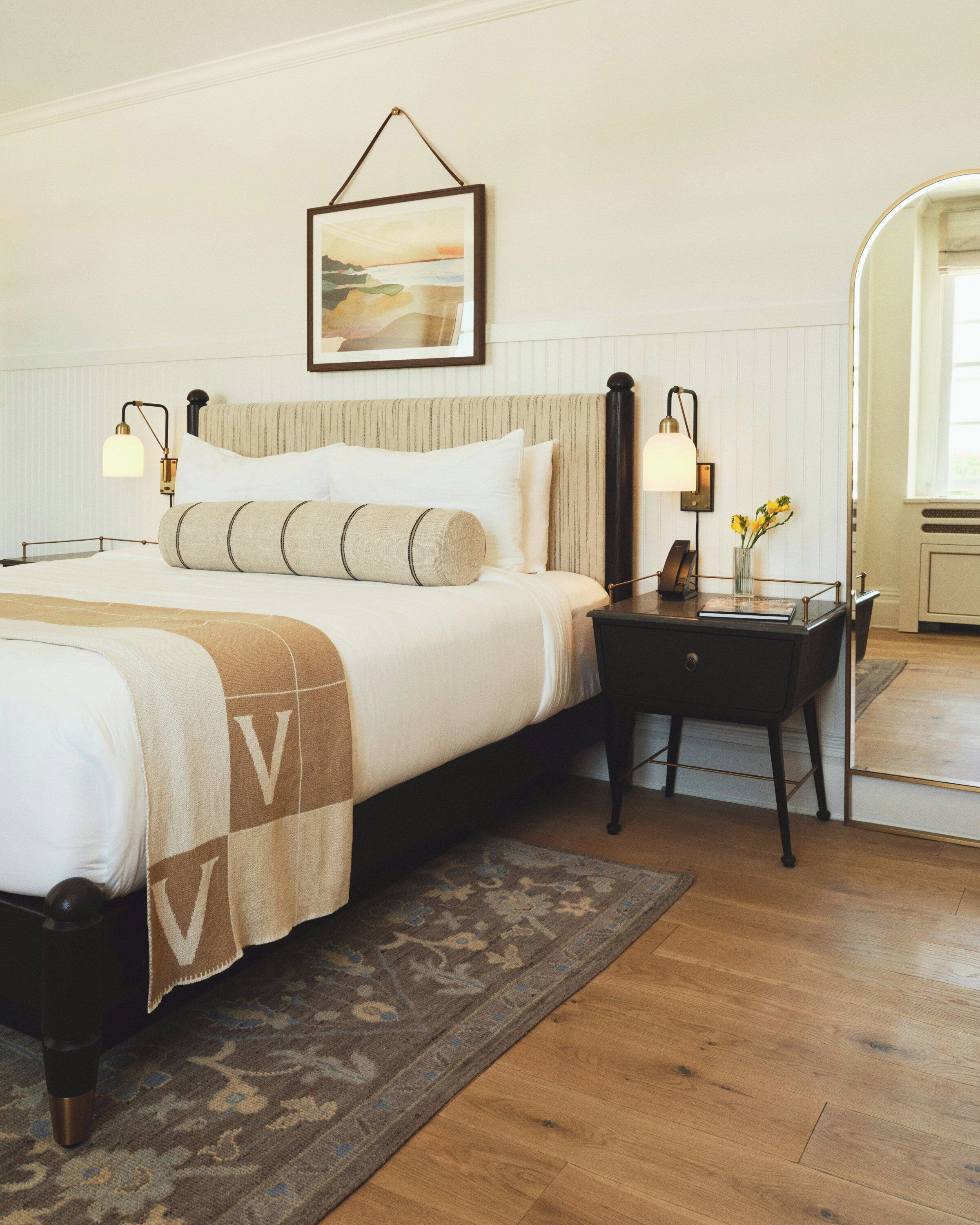 A neatly arranged bedroom with a bed, bedside lamps, a desk, and a framed picture on the wall.