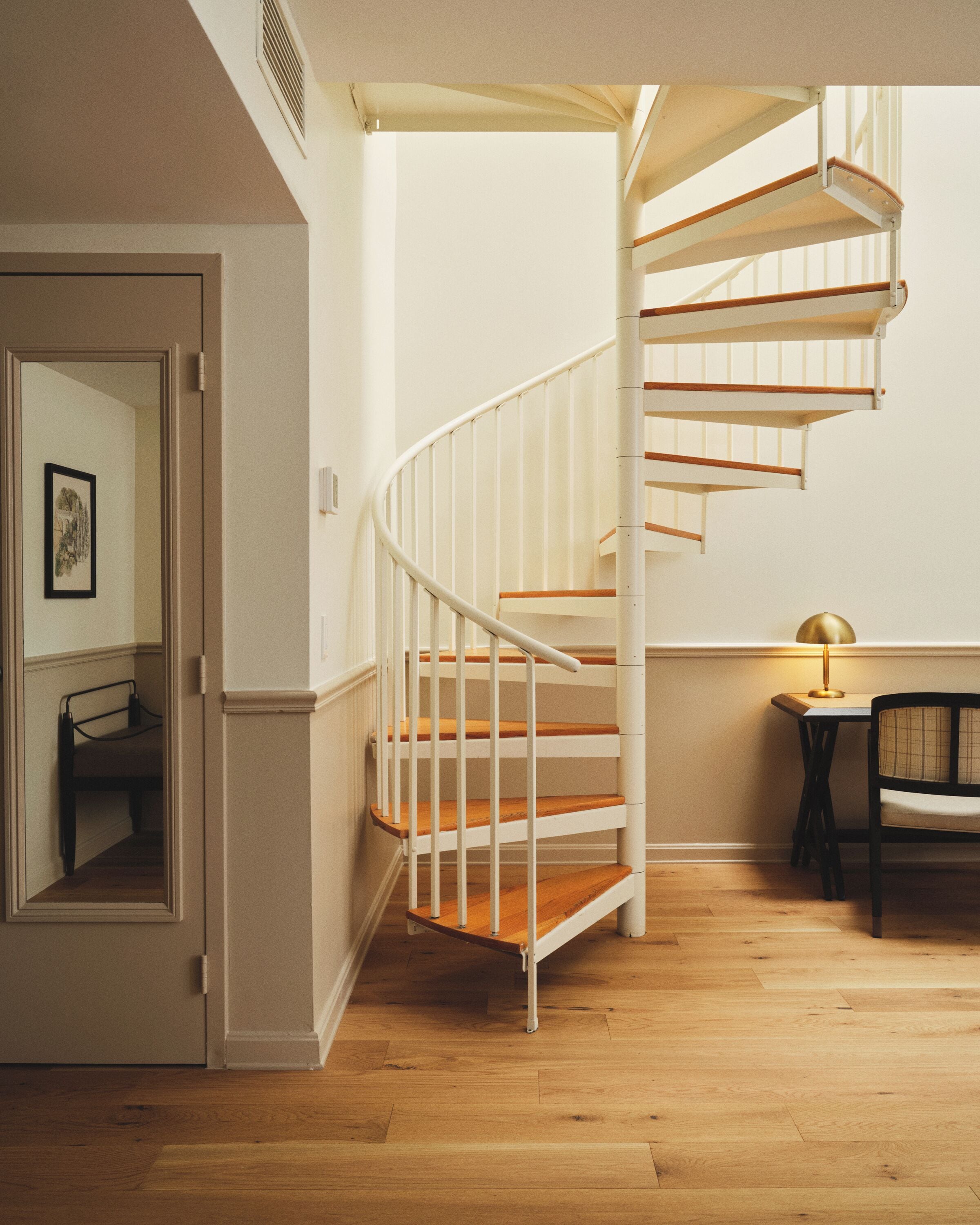 Un escalier en colimaçon avec des marches en bois et une rampe blanche, une petite table avec une lampe et une chaise dans une pièce dont le sol est en bois et qui possède une porte ornée d’un tableau encadré.