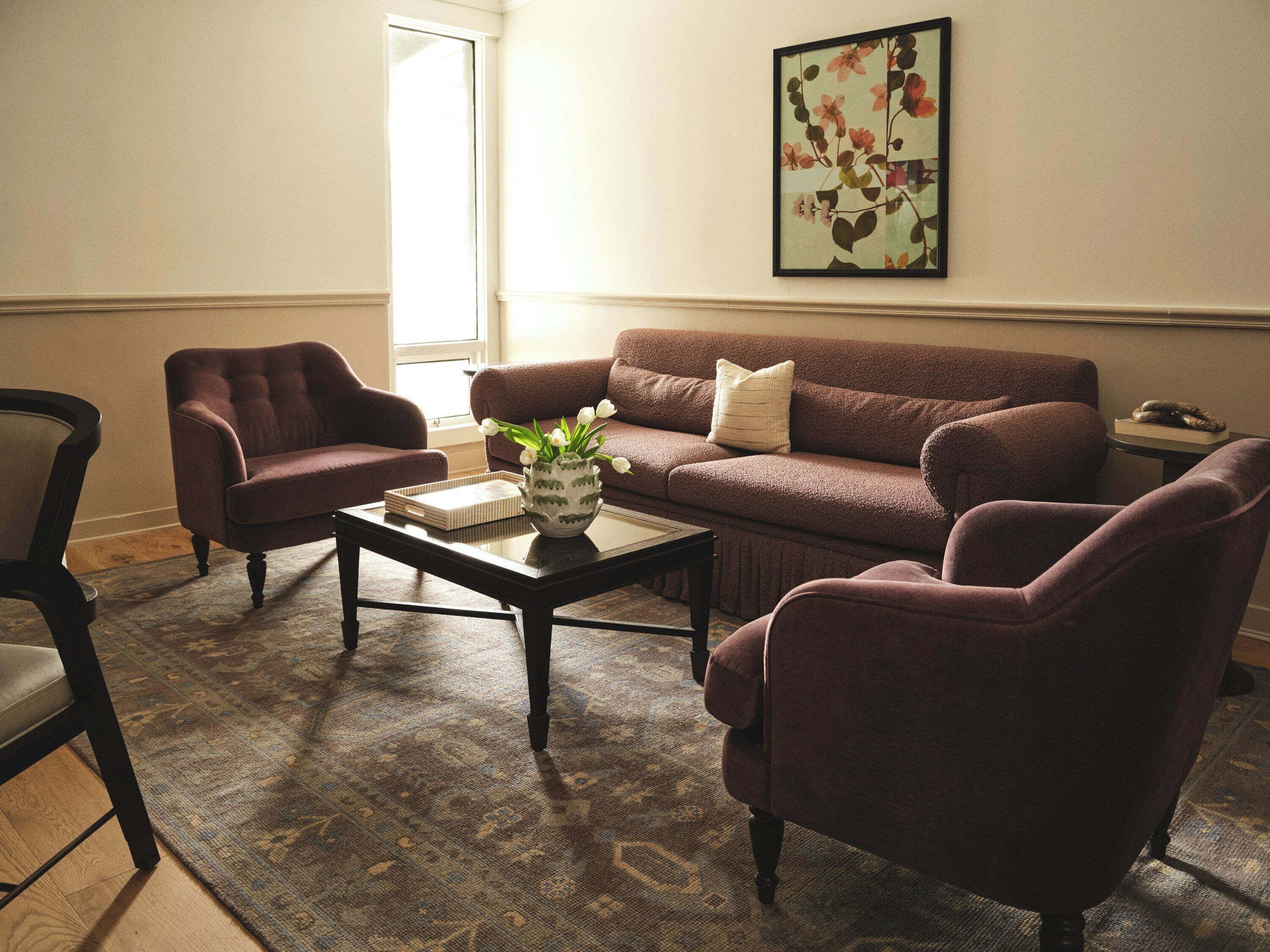 A living room with a brown sofa, two armchairs, a coffee table with a vase of flowers, and a framed picture on the wall.