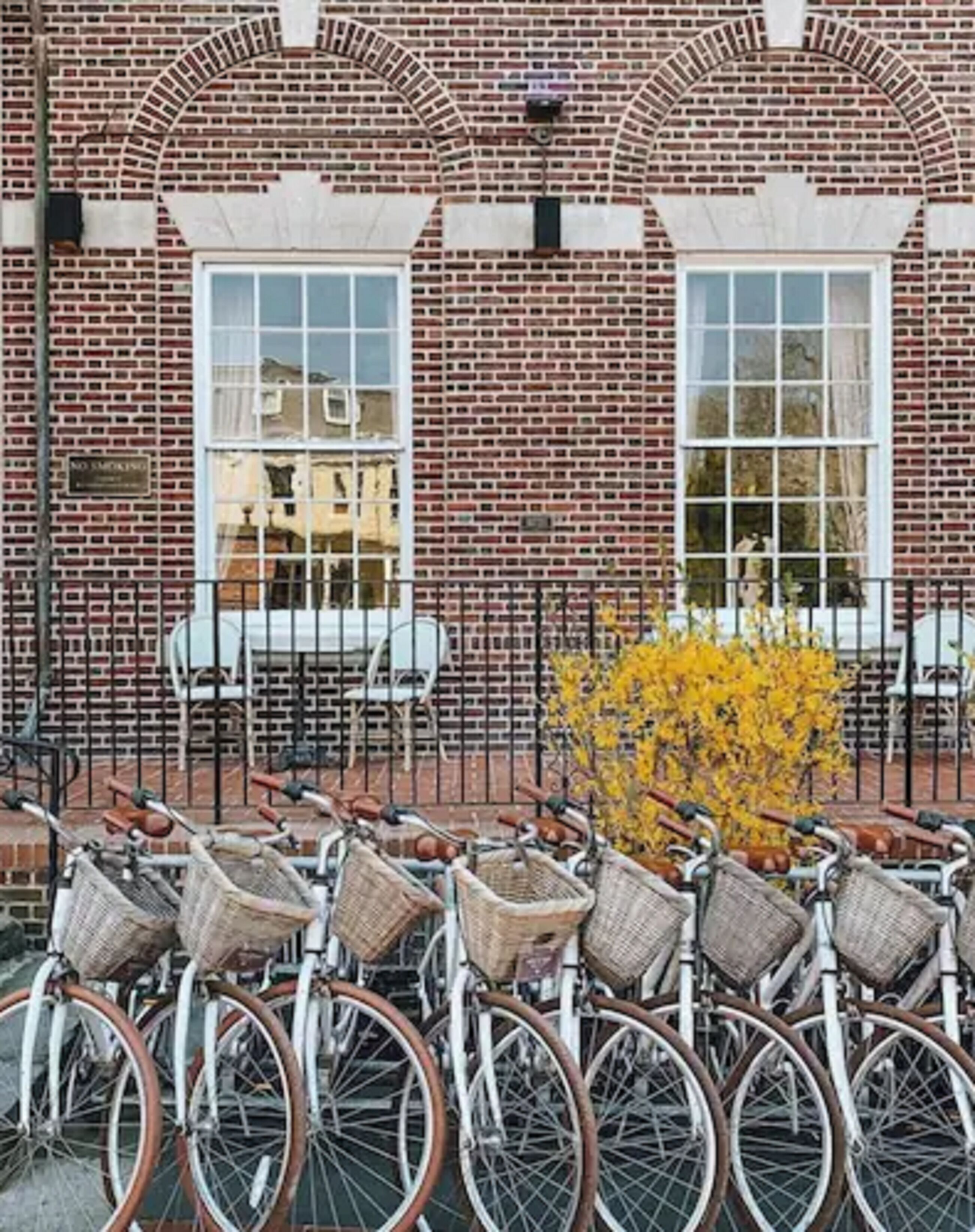 A row of bicycles with baskets parked in front of a brick building with arched windows.
