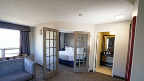 Une chambre d’hôtel avec un canapé gris, un lit avec du linge de lit blanc, une salle de bain avec un miroir et un lavabo, et une porte donnant sur une autre pièce.
