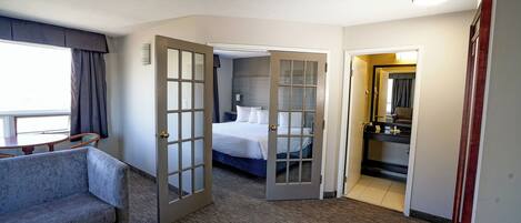 A hotel room with a gray sofa, a bed with white linens, a bathroom with a mirror and sink, and a door leading to another room.