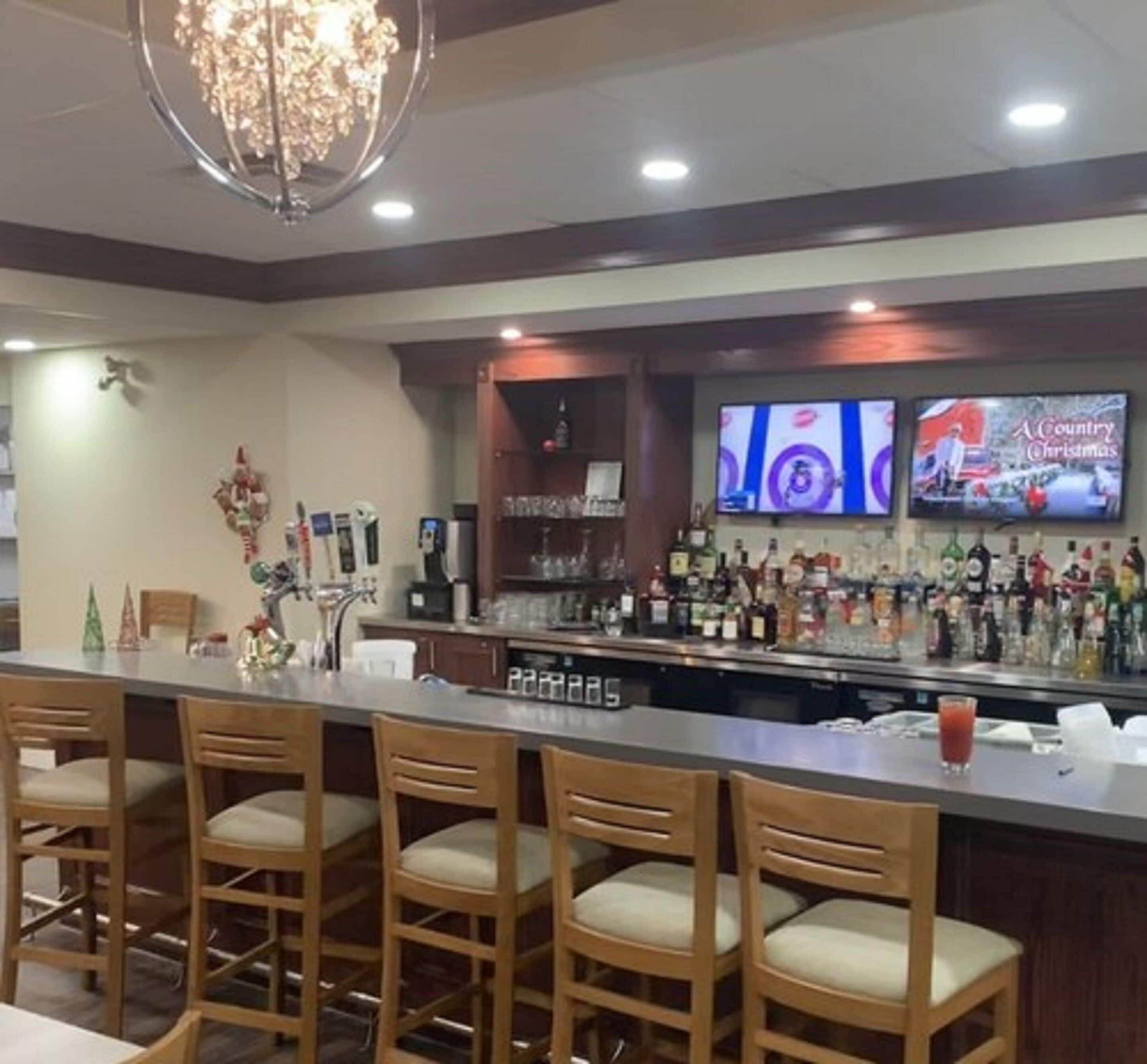 A bar area with wooden bar stools, a counter with a variety of liquor bottles, and a television screen displaying a Christmas program.