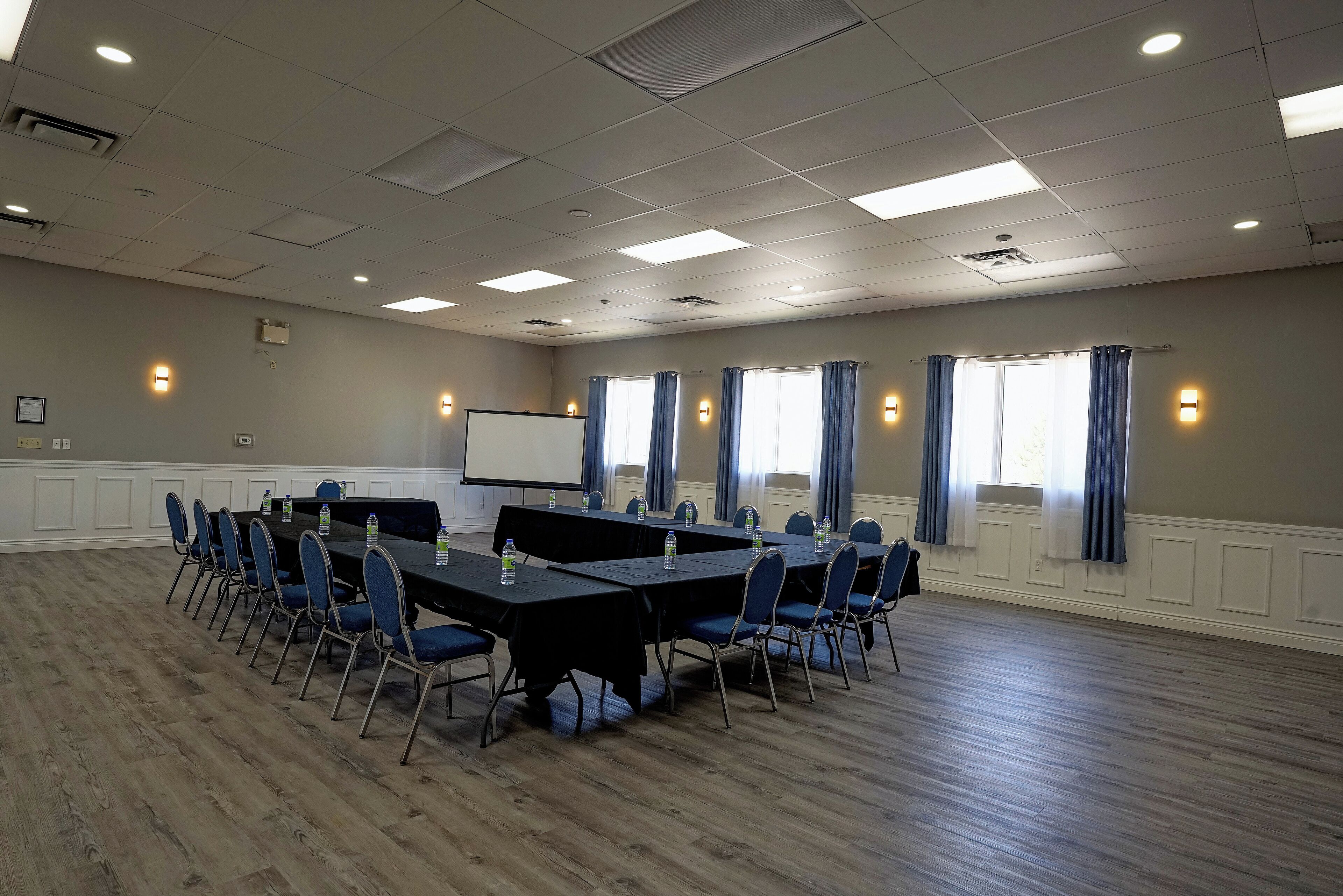A conference room with a long table, chairs, a projector screen, and windows with curtains.