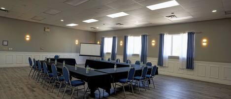 Un salón de conferencias con una larga mesa, sillas, una pantalla de proyector y ventanas con cortinas.