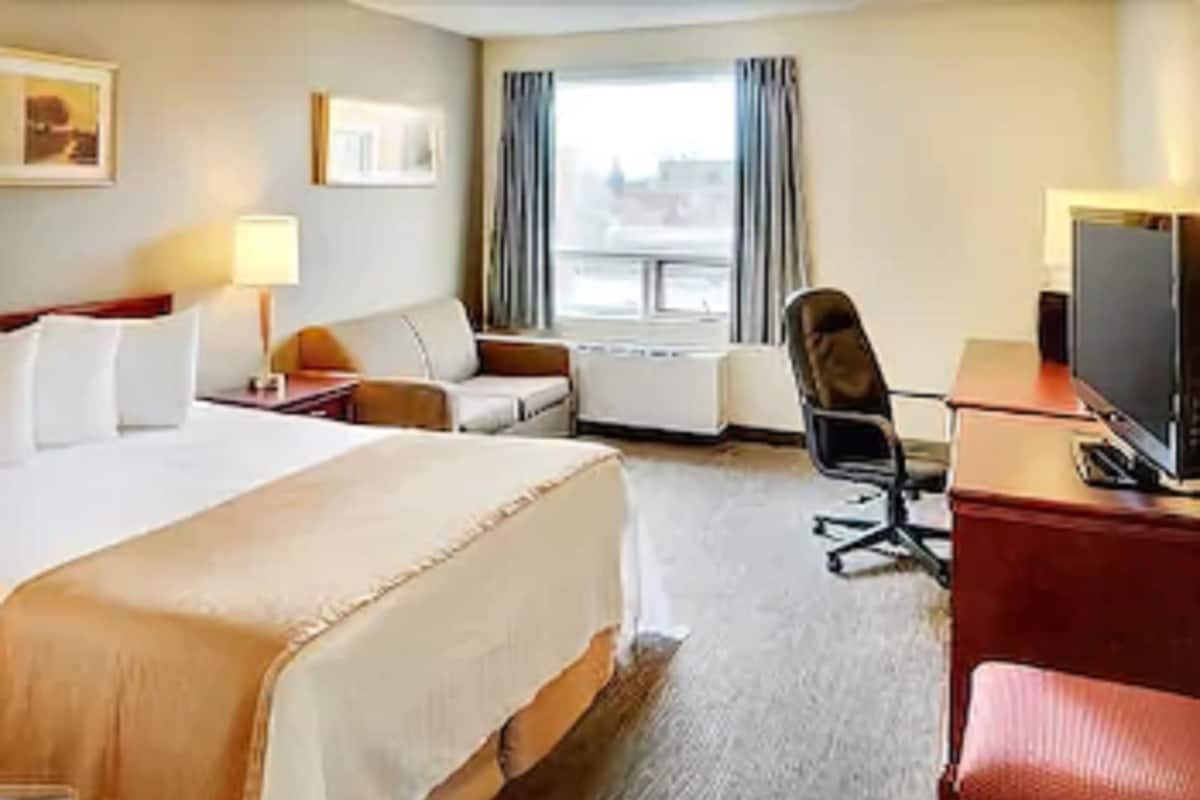 Ein Hotelzimmer mit einem großen Bett, einem Schreibtisch mit Computer, einem Sessel, einem Fernseher und einem Fenster mit Vorhängen.