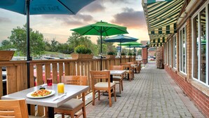 Une terrasse extérieure avec des tables et des chaises en bois, des parasols verts et des murs en briques.