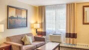 Quarto de hotel com sofá, pequena mesa, luminária, pintura, janela com cortinas e ar condicionado.