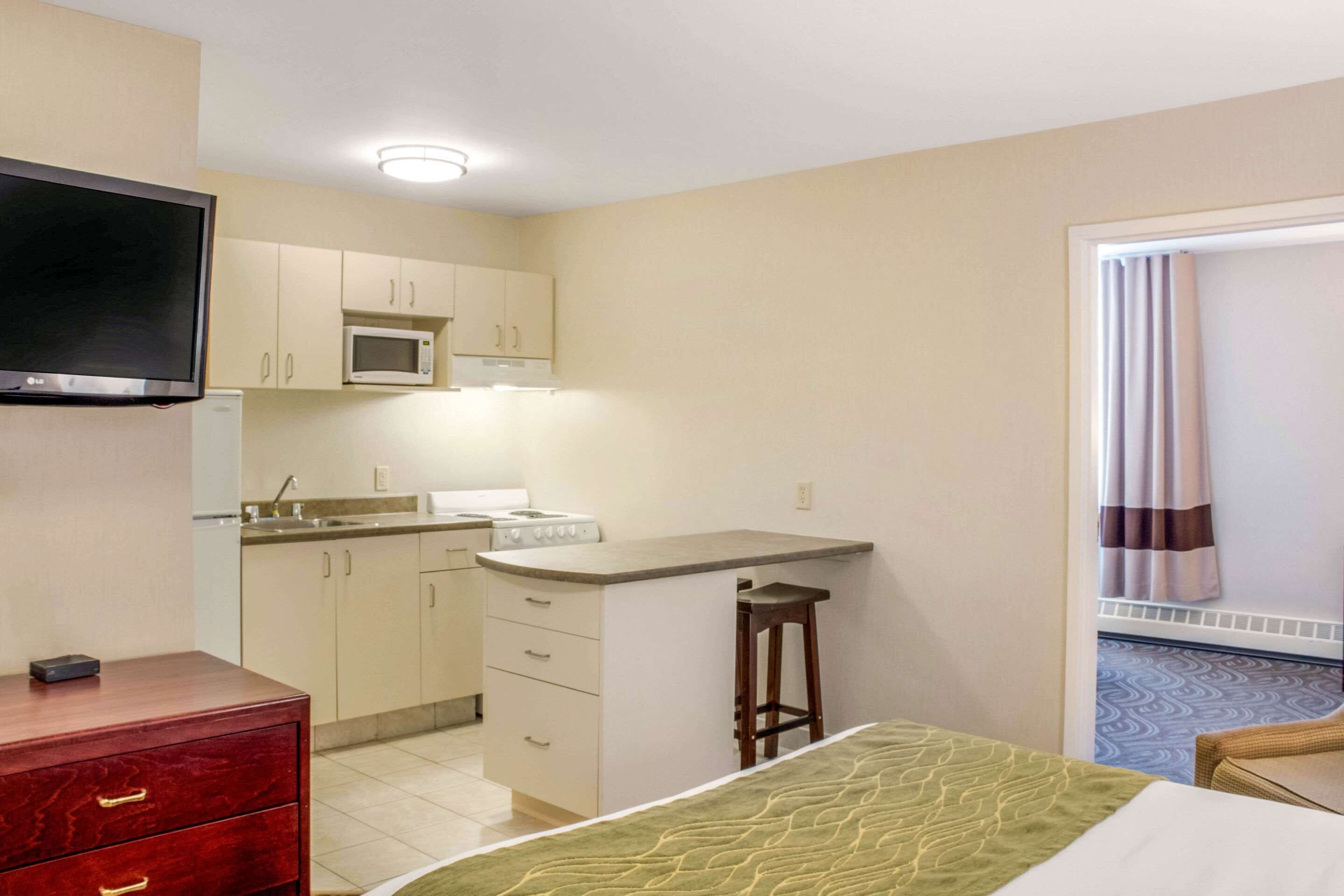Een compacte hotelkamer met een kitchenette, een bed met een groene plaid, een houten dressoir en een aan de muur bevestigde televisie.