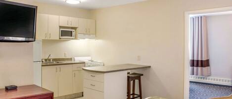 A compact hotel room with a kitchenette, a bed with a green bedspread, a wooden dresser, and a television mounted on the wall.