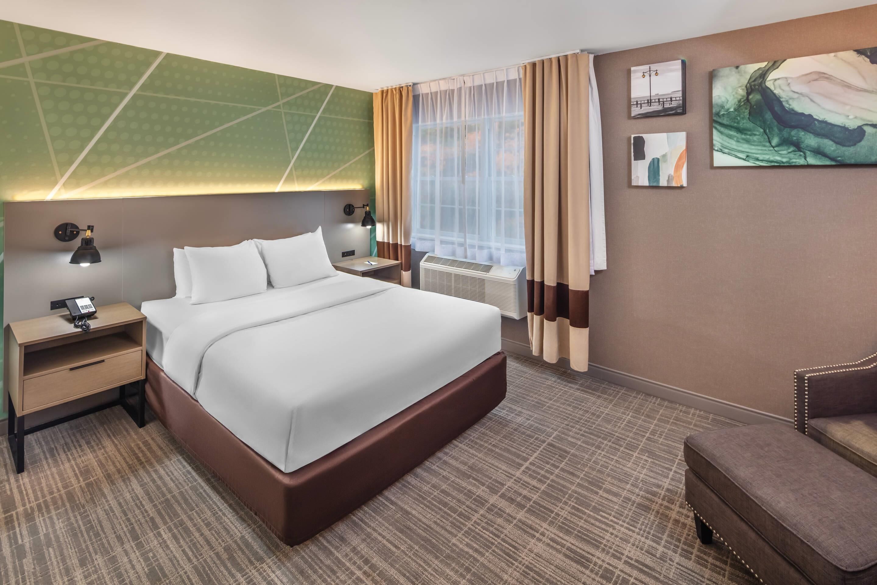 A hotel room with a large bed, a nightstand, a desk, a chair, a window with curtains, and artwork on the walls.