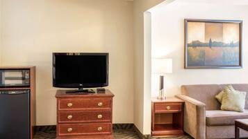 Quarto de hotel com televisão de tela plana sobre uma cômoda de madeira, micro-ondas, mesa pequena com abajur, sofá e um quadro na parede.