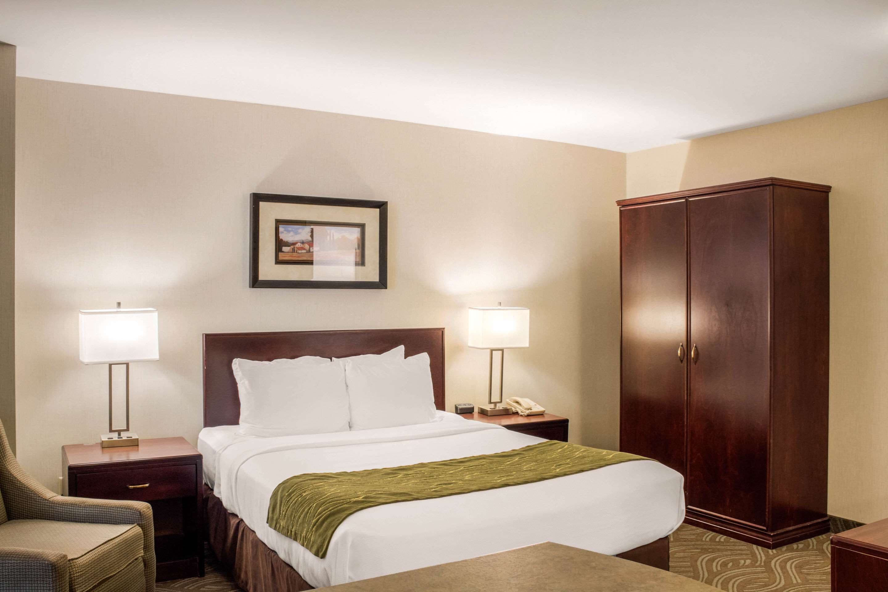 A hotel room with a bed, two bedside lamps, a nightstand, a chair, a wardrobe, and a framed picture on the wall.