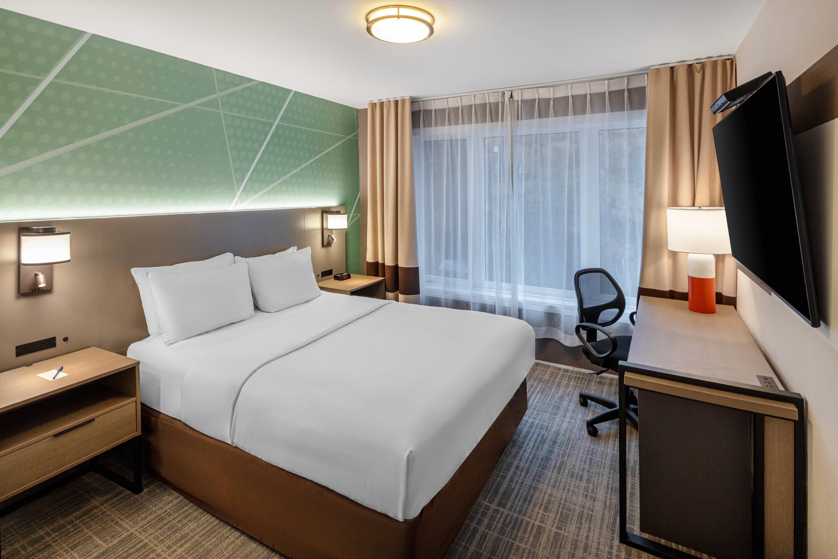 A hotel room with a large bed, a desk with a chair, a TV, and a window with curtains.