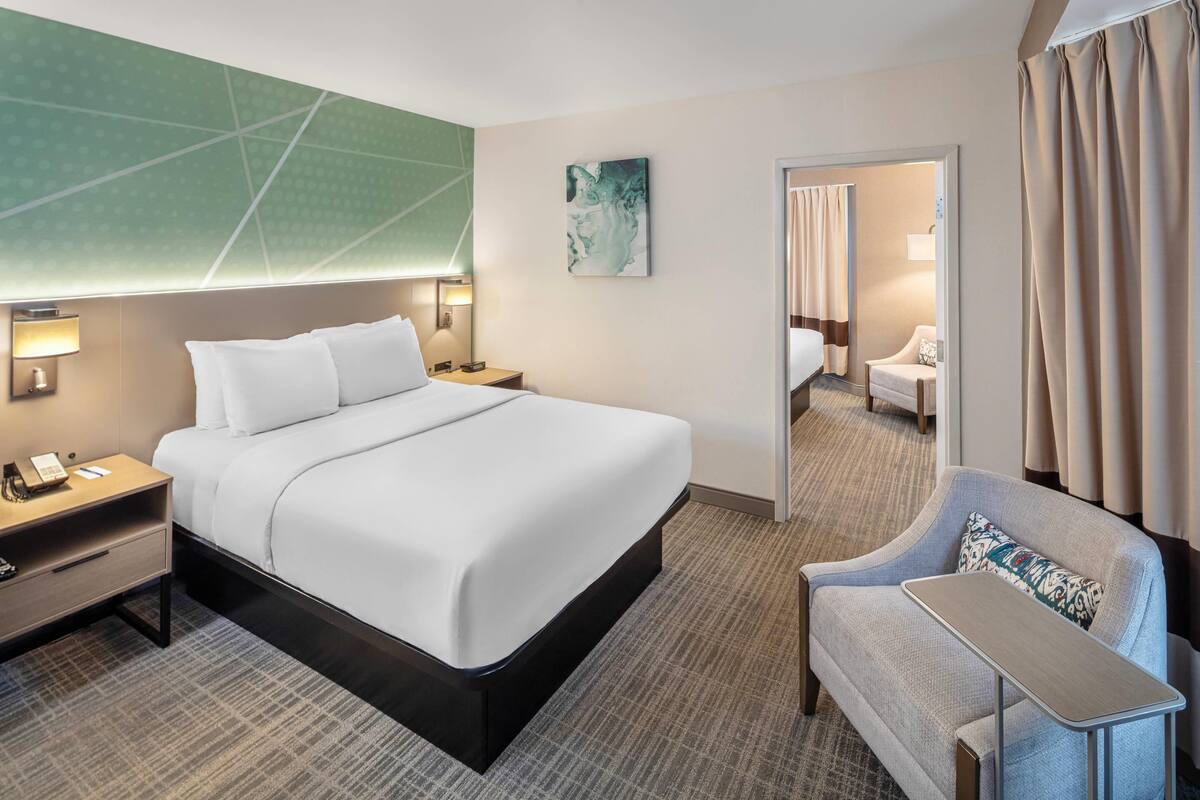 Une chambre d’hôtel avec un grand lit, des tables de chevet avec des lampes, un petit fauteuil et un miroir.