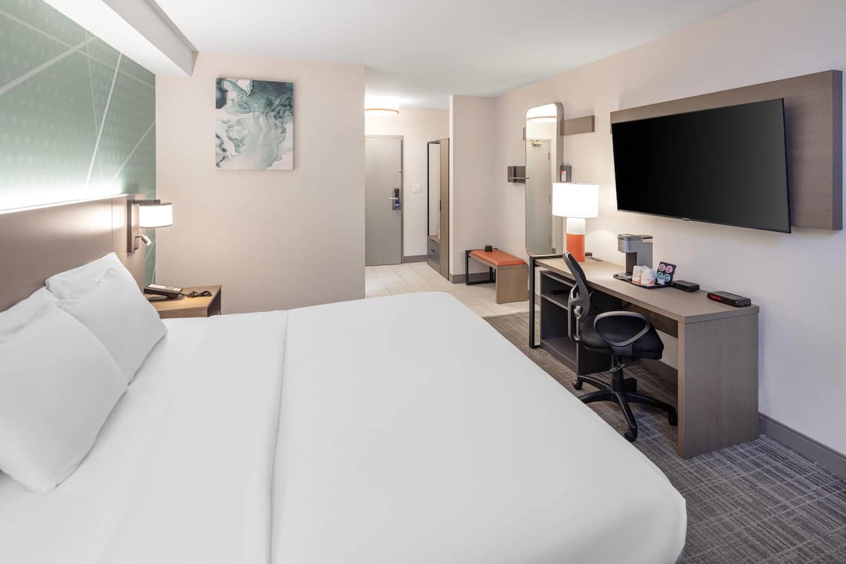 Une chambre d’hôtel avec un grand lit, un bureau avec une chaise, un téléviseur à écran plat fixé au mur et une salle de bain visible à travers une porte ouverte.