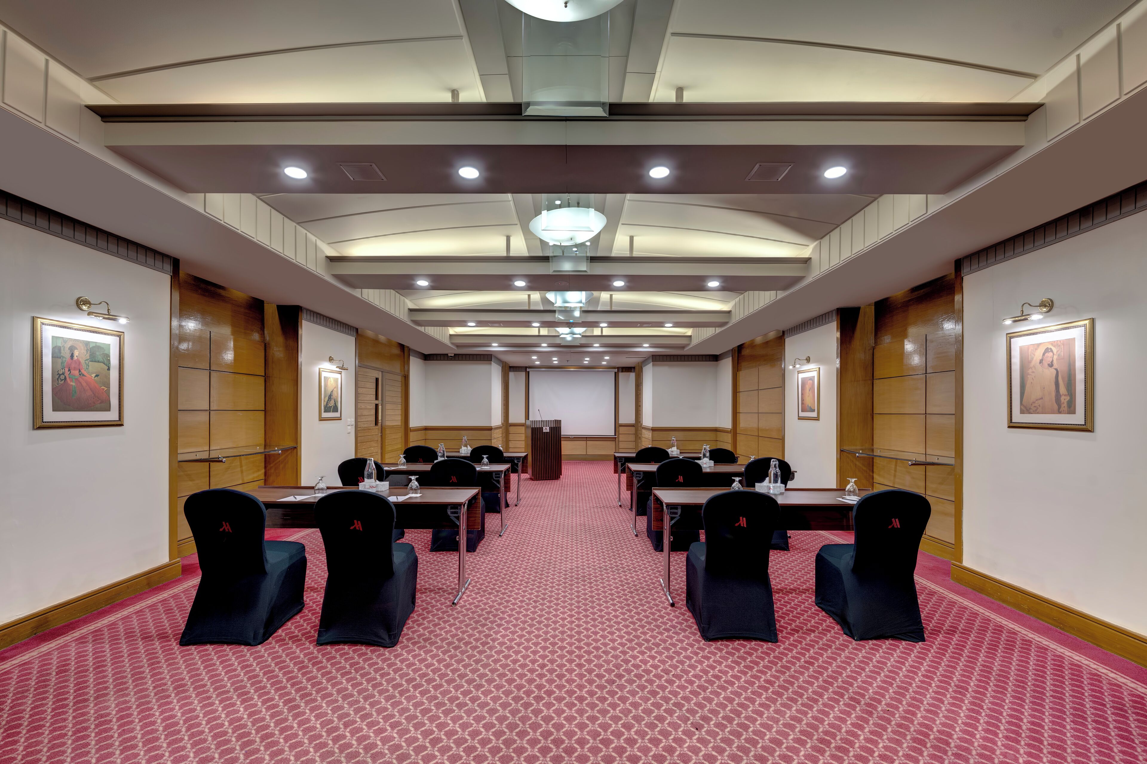 A conference room with a red patterned carpet, multiple tables with chairs, a podium, and framed pictures on the walls.
