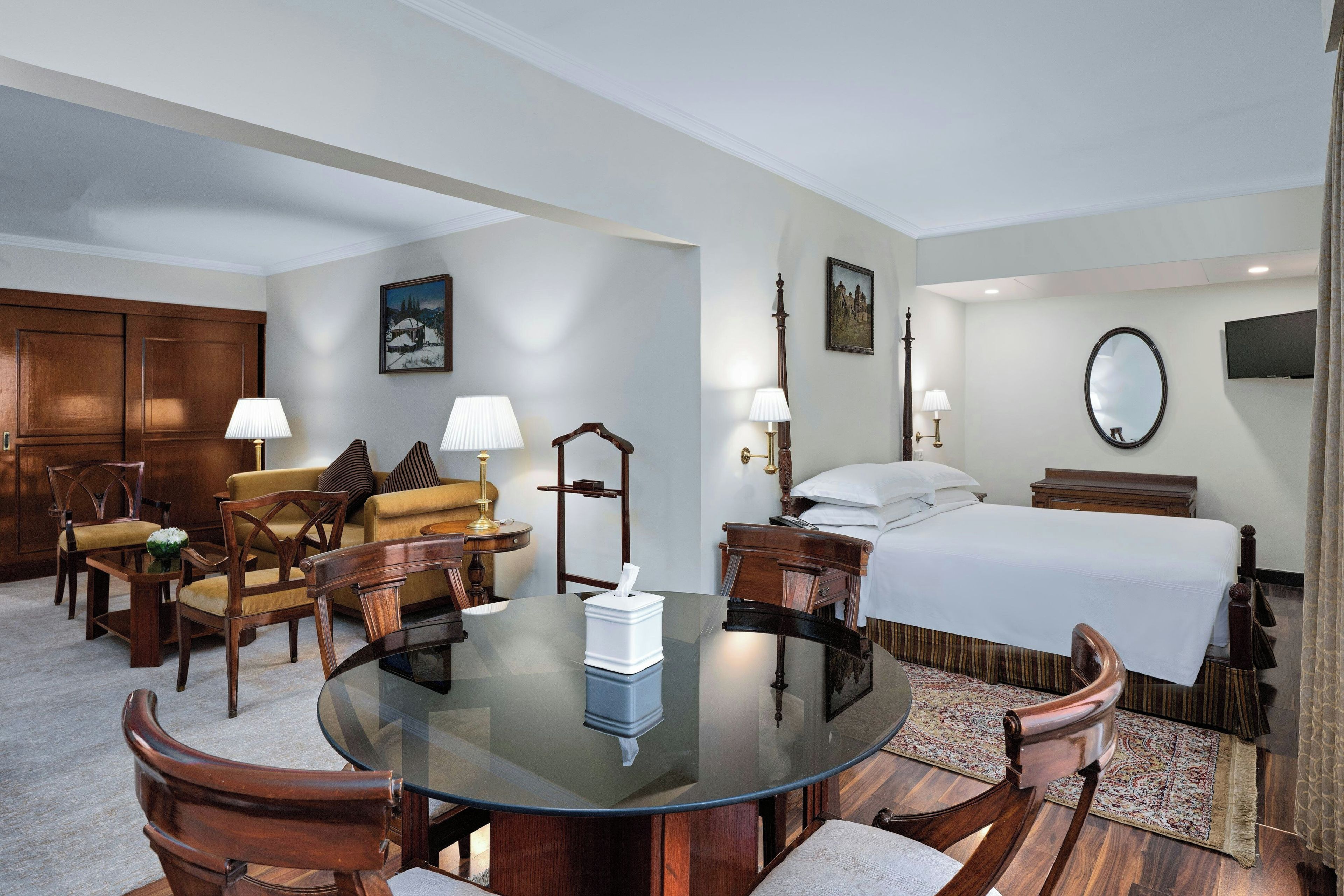 Une chambre d’hôtel avec une table ronde en verre, des chaises en bois, un canapé, un lit, une télévision et un miroir.