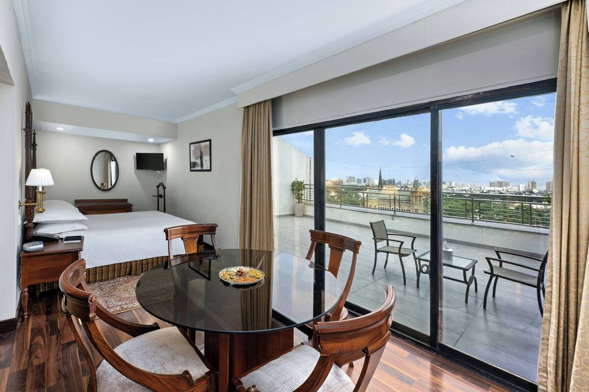 A hotel room with a balcony, a bed, a dining table with chairs, a TV, and a lamp.