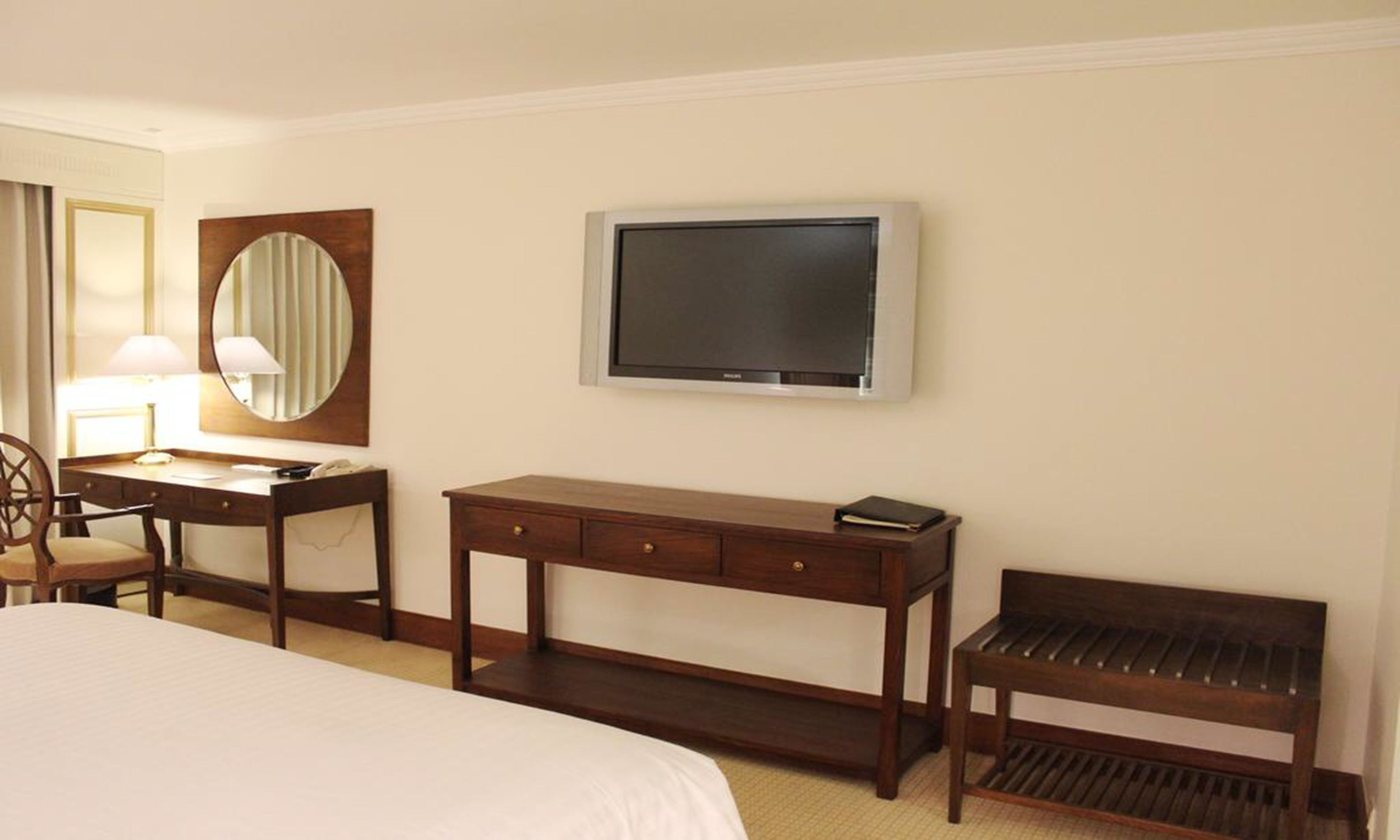 Een hotelkamer met een bed, een bureau met lamp, een televisie, een spiegel en een bankje.