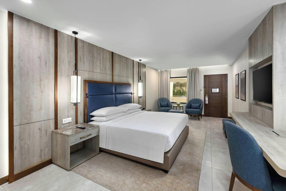 A modern hotel room with a bed, desk, chairs, and a TV.