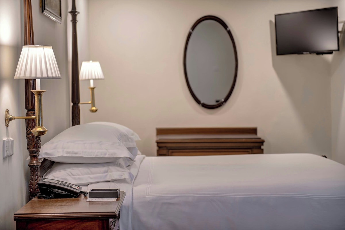 Habitación de hotel con cama, lámparas de noche, teléfono, espejo y televisor montado en la pared.
