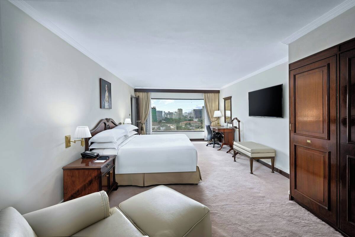 A hotel room with a large bed, a desk, a chair, a TV, and a view of the city.