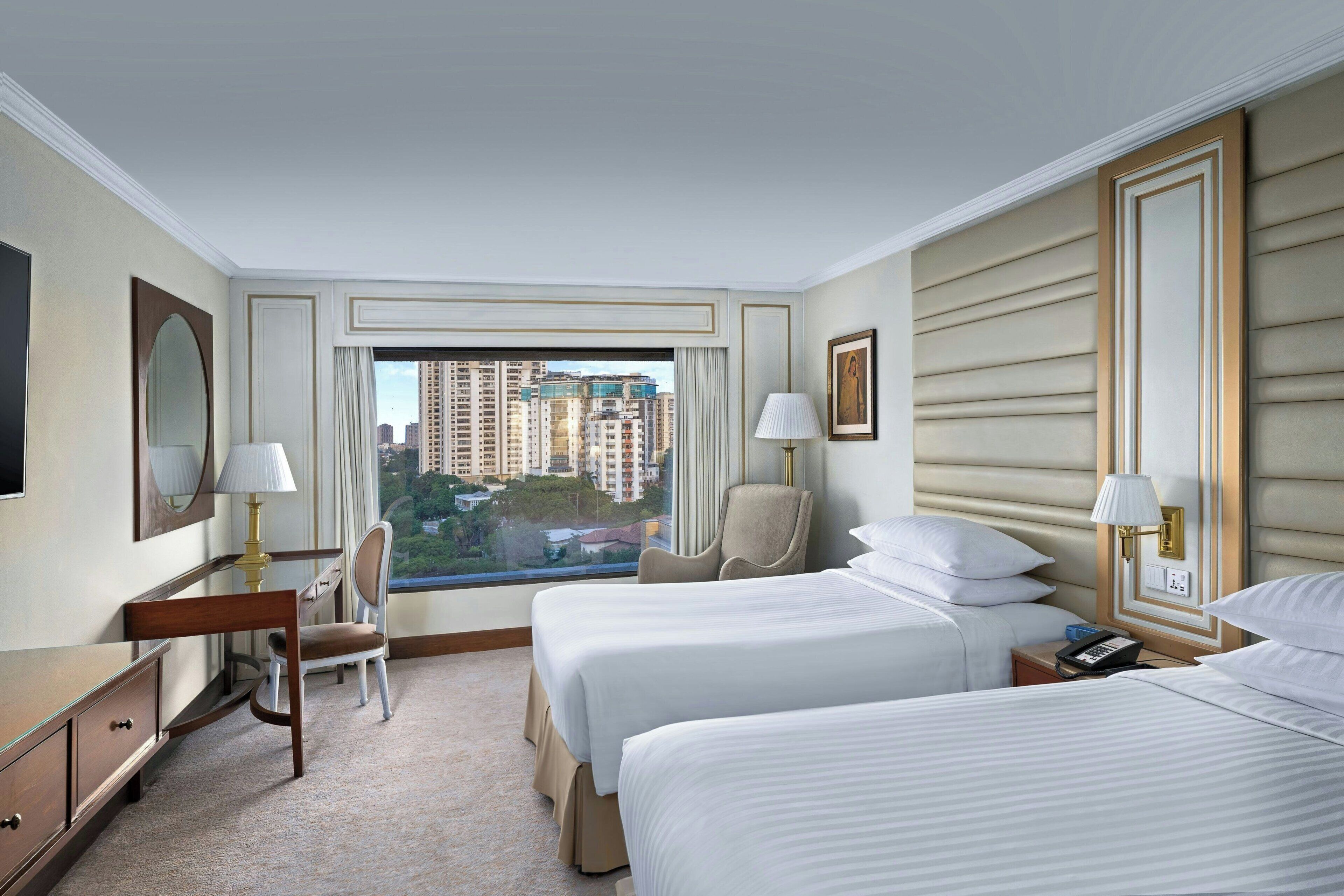 A hotel room with two beds, a desk, a chair, and a large window offering a city view.