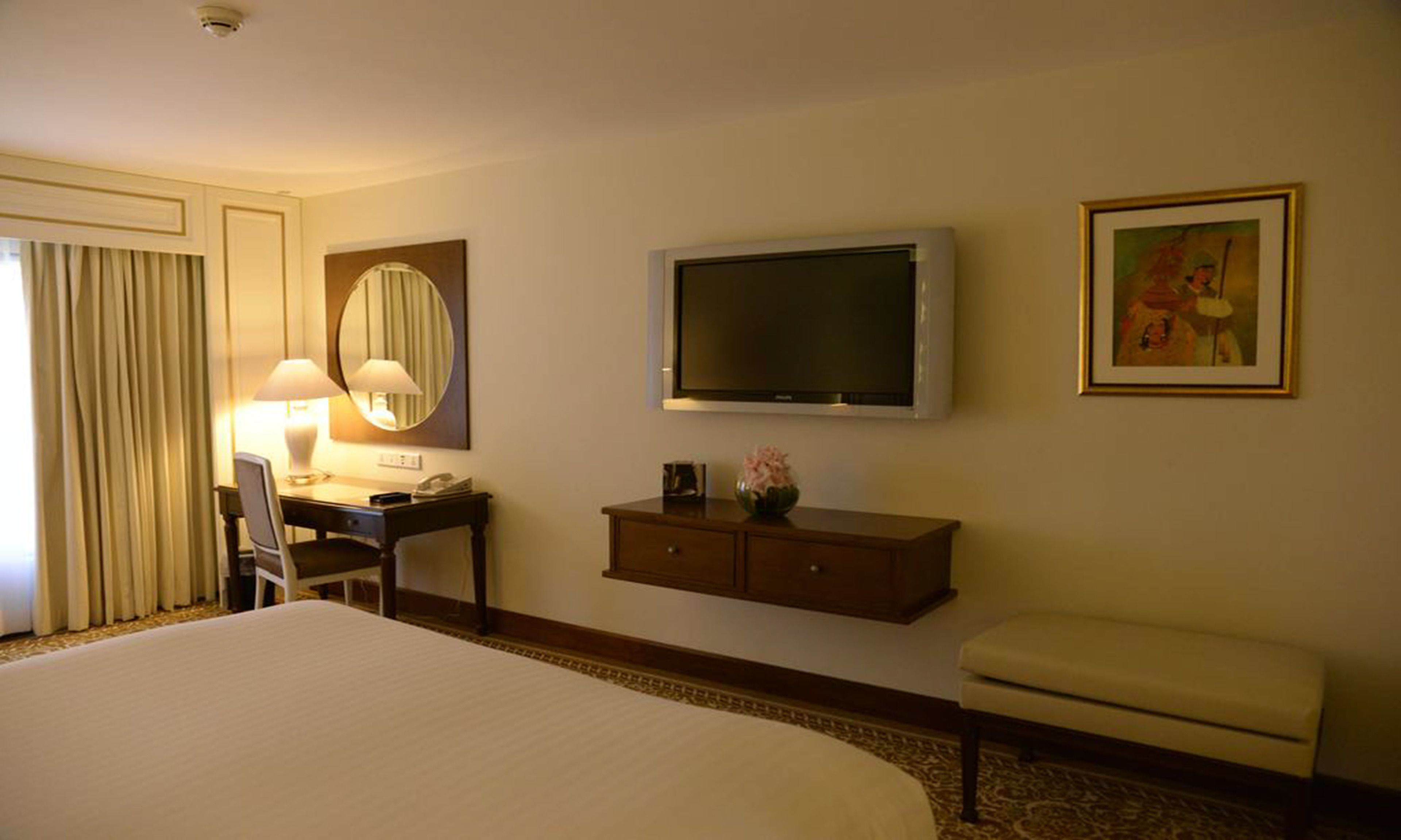 A hotel room with a large bed, a desk with a lamp, a television, a small cabinet, and a painting on the wall.