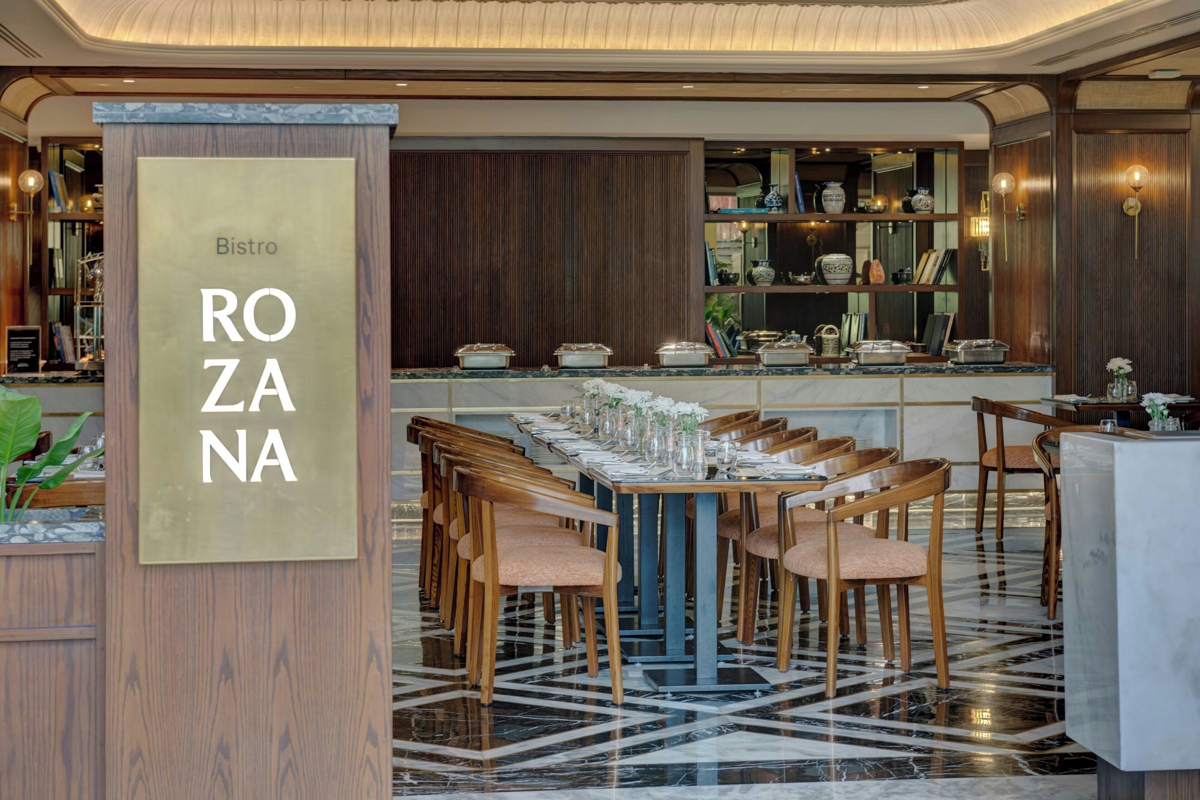 Un restaurant doté d’un sol en marbre, de chaises en bois, d’une longue table dressée pour le repas et d’une enseigne indiquant « Bistro ROZANA ».