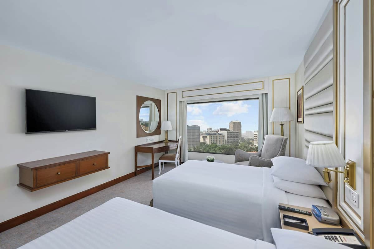 A hotel room with two beds, a TV, a desk, a chair, a window with a city view, and a lamp.