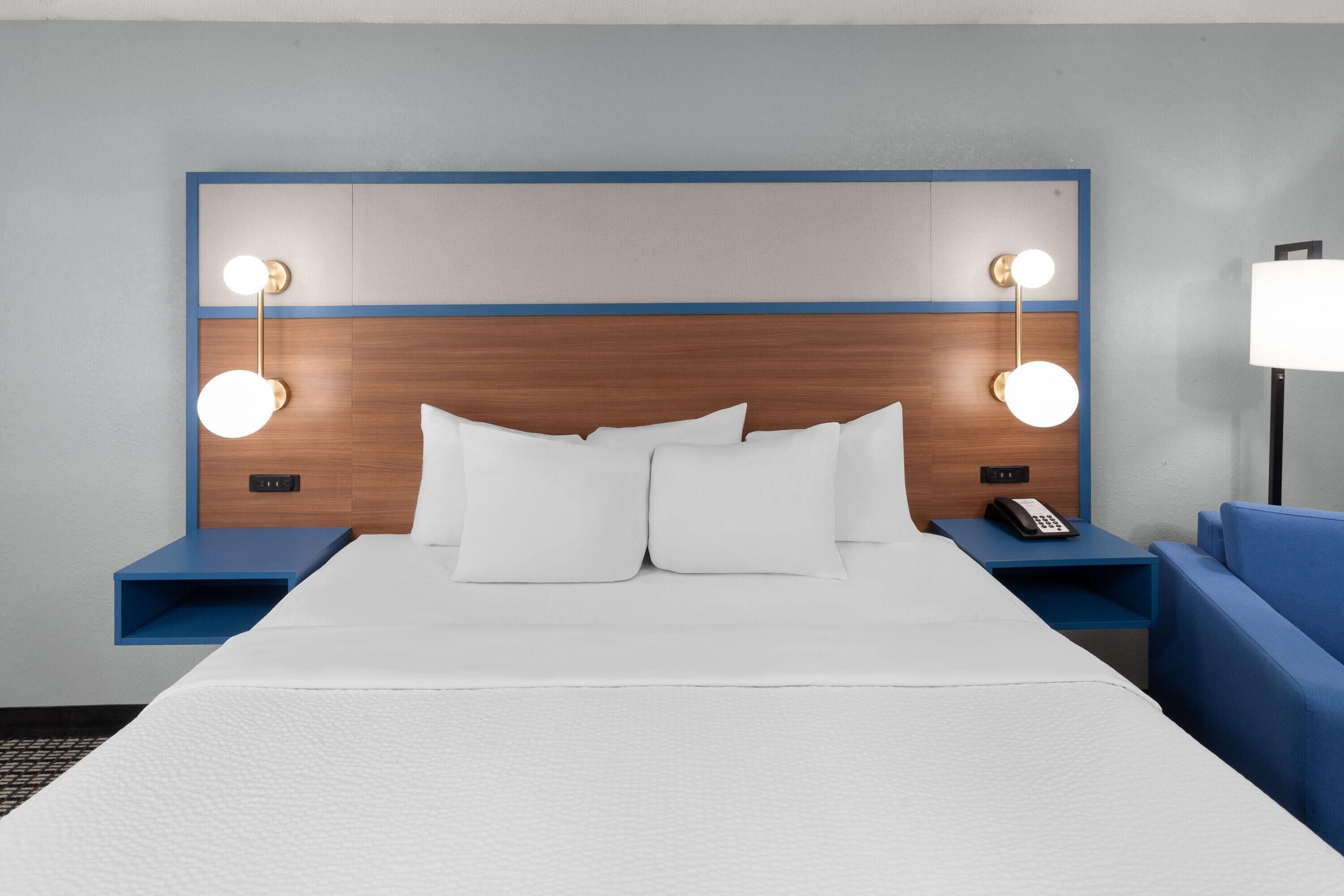 A hotel room with a large bed, two bedside tables, a nightstand, a blue sofa, and a wall-mounted lamp.