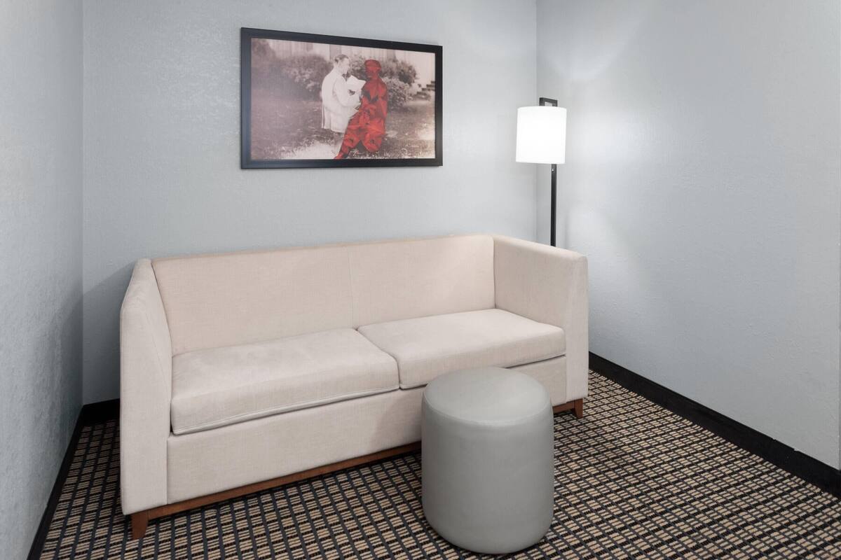 A room with a beige sofa, a round ottoman, a patterned floor, a framed picture on the wall, and a floor lamp.